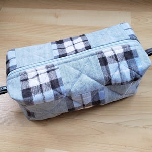 May include: A rectangular, quilted fabric pouch with a light blue plaid pattern. The pouch has a light blue zip and small fabric loops on each end. The plaid design incorporates white and dark grey squares.