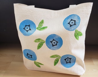 May include: A white canvas tote bag with blue and green painted blueberries. The blueberries are stylized and have a five-pointed star in the center.