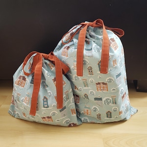 May include: Two drawstring bags with a light blue background and a repeating pattern of houses in various colours. The bags have rust-coloured handles and drawstrings. The bags are made of fabric.