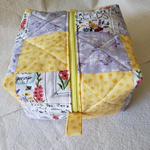 Peut inclure: Une pochette en tissu matelassé avec une fermeture éclair jaune et une tirette. La pochette est composée de carrés de patchwork en pois jaunes, fleurs de lavande et imprimés floraux. Le texte "Seeds" et "Plant these" est visible.