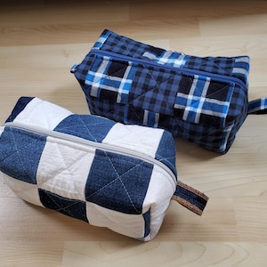May include: Two quilted fabric pouches. One is white and blue checkered, the other is blue and black plaid. Both have a zip closure and a small fabric loop on the side. The pouches are rectangular and appear to be handmade.
