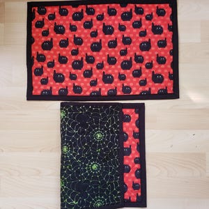 May include: A rectangular quilted mat featuring a repeating pattern of small black cats on a bright red background with a black border. Below it, a folded quilted mat displays a black spiderweb pattern with green accents on one side.