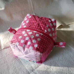 May include: A handmade, quilted fabric cube-shaped pouch. The pouch features a pink and white polka dot pattern, a pink gingham pattern, and a pink zip. It has small pink fabric loops on each side. The pouch is likely used for storage.