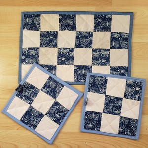 May include: A quilted kitchen set comprising a rectangular placemat and two square pot holders. The set features a checkerboard pattern with alternating white and navy blue squares. The navy squares are decorated with a butterfly print, and the edges have a light blue border.