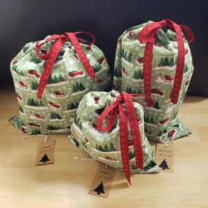 Peut inclure: Trois sacs cadeaux en tissu avec cordon de serrage, en tailles petite, moyenne et grande. Les sacs présentent un fond vert clair avec un motif de camions rouges, de traîneaux et de sapins. Chaque sac a un ruban à carreaux rouges.