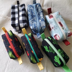 Peut inclure: Plusieurs pochettes rectangulaires zippées avec différents motifs. Les motifs incluent des carreaux noirs et blancs, des papillons bleus, des maisons d'hiver, des cactus verts et des piments. Chaque pochette a une fermeture éclair et une tirette en tissu assorties.