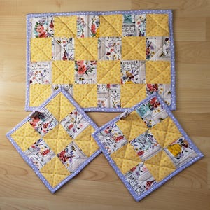 May include: A quilted placemat and two matching pot holders. The set features a patchwork design with yellow squares and floral print squares. The edges are trimmed with a light blue polka dot fabric.