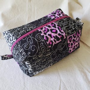 May include: A rectangular cosmetic bag with a pink zip. The bag features a patchwork design with black fabric adorned with white script and swirls, and pink and black leopard print sections. A small fabric loop is attached to the side.