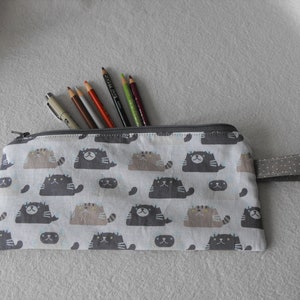 May include: A grey zipped pencil case with a white background and a repeating pattern of cartoon cats. The case has a grey fabric handle with white polka dots. The case is partially open and has several coloured pencils inside.
