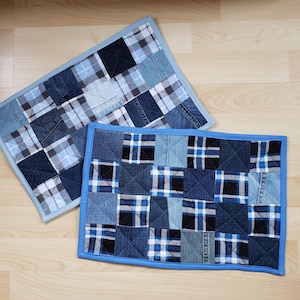 May include: Two denim patchwork placemats. One is rectangular with a light blue border and a patchwork design of blue and white plaid and solid denim squares. The other is square with a dark blue border and a patchwork design of blue, black, and white plaid and solid denim squares.