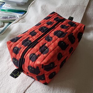 May include: A quilted rectangular pouch with a black zipper and pull tab. The red fabric is patterned with black cat silhouettes with green eyes. The pouch is likely used for storage or travel.