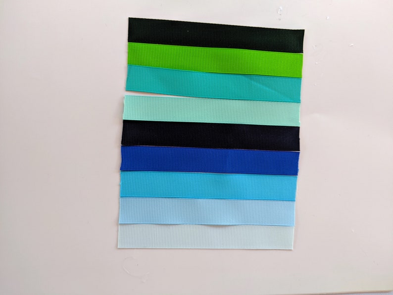 May include: A collection of eight ribbon samples in various shades of blue, green, and black. The ribbons are arranged vertically in a row, with the darkest color at the top and the lightest color at the bottom.