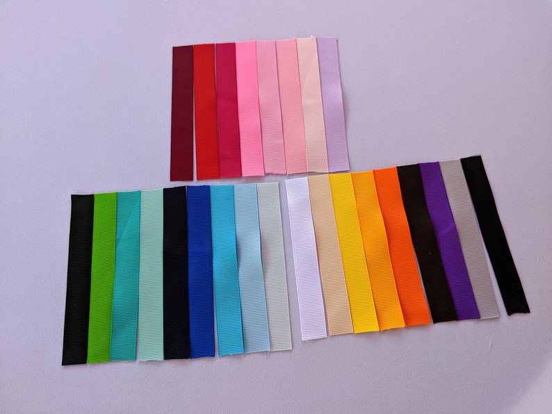 May include: Assortment of 20 different colored ribbon spools. The ribbons are all the same width and are arranged in two rows. The top row has 8 ribbons in shades of red, pink, and purple. The bottom row has 12 ribbons in shades of green, blue, black, white, yellow, and orange.