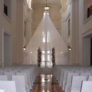 May include: A white fabric canopy hangs over a wedding ceremony setup in a large, white room with white chairs arranged in rows. The room has tall windows and white walls.