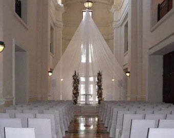May include: A white fabric canopy hangs over a wedding ceremony setup in a large, white room with white chairs arranged in rows. The room has tall windows and white walls.