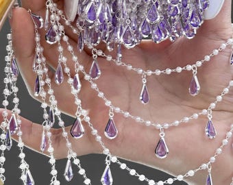 May include: Close-up of decorative beaded trim with purple teardrop-shaped pendants and small white beads. The trim is on a roll, with strands of the trim cascading down. The trim is likely used for embellishing clothing or crafts.