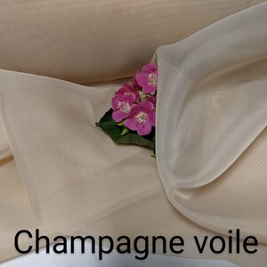 May include: Champagne voile fabric, a sheer, lightweight material with a delicate texture. The fabric is a soft, pale champagne color and is draped over a white surface. A cluster of pink flowers is placed on the fabric.