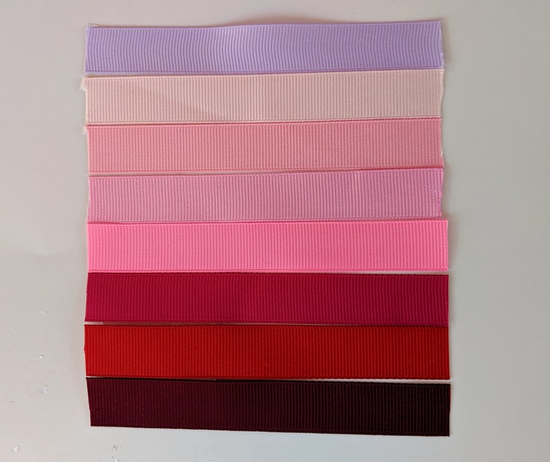 May include: A collection of eight different colored ribbon spools. The colors range from light purple to dark burgundy. The ribbon is a satin material with a slight sheen.