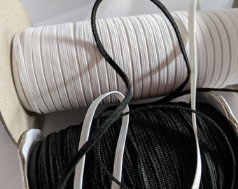 Pode incluir: Close-up de rolos de cordão elástico branco e preto. O elástico branco está num rolo grande, o preto num mais pequeno. Um pedaço de elástico branco e preto é visível.