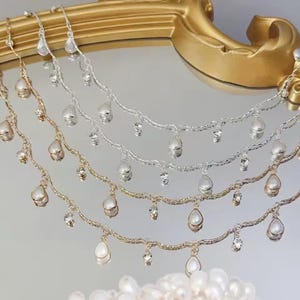 May include: Close-up of several delicate necklaces. The necklaces are gold and silver, each featuring teardrop-shaped pearls and small, sparkling crystals. The jewelry is displayed on a reflective surface.