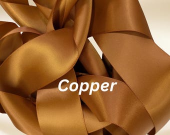 Puede incluir: Primer plano de cinta de raso color cobre, dispuesta en un patrón arremolinado. La cinta tiene un acabado liso y brillante, que refleja la luz. La palabra "Copper" está impresa en blanco en la parte inferior de la imagen.
