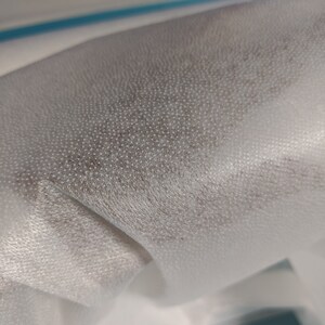 May include: Close-up of white non-woven fabric with a textured surface. The fabric is folded and has a slight sheen.
