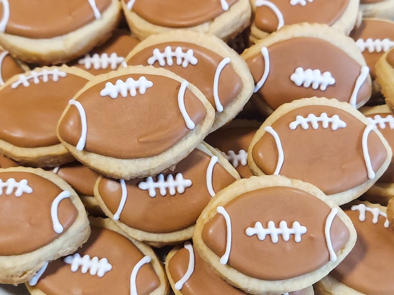 May include: A close-up of a pile of brown and white football-shaped cookies. The cookies are decorated with white icing and have a brown glaze. The cookies are arranged in a random pattern and are all facing the camera.