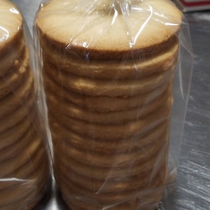 May include: Two bags of round, light brown cookies stacked high. The cookies are in clear plastic bags.