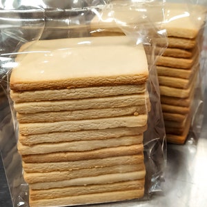 May include: A stack of plain, rectangular cookies in a clear plastic bag. The cookies are light brown and have a slightly rough texture.