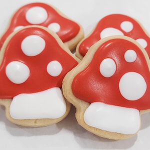 May include: Four mushroom-shaped cookies with red icing and white polka dots. The cookies have a white base and a light brown edge. The cookies are arranged on a white surface.