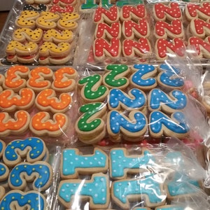 May include: Assortment of decorated sugar cookies in various colors and designs, including polka dots, stripes, and numbers. The cookies are arranged in clear plastic bags on a metal surface.