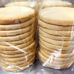 May include: Two stacks of plain round cookies wrapped in clear plastic bags. The cookies are light brown and stacked neatly.
