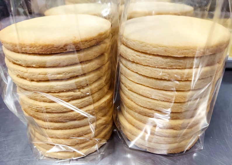May include: Two stacks of plain round cookies wrapped in clear plastic bags. The cookies are light brown and stacked neatly.