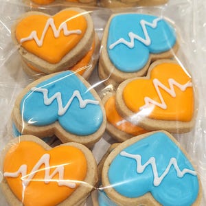 May include: Heart-shaped cookies with orange and blue frosting, each decorated with a white electrocardiogram design. The cookies are packaged in clear plastic, showcasing the baked goods.