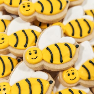 May include: A close-up of a pile of yellow and black bee-shaped sugar cookies with white icing wings. The cookies have black lines to represent the bee's stripes and a smiling face.