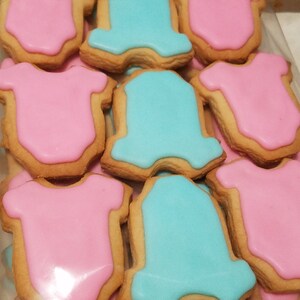 May include: A close-up of a variety of sugar cookies decorated with blue and pink icing in the shape of onesies. The cookies are arranged in a plastic bag.
