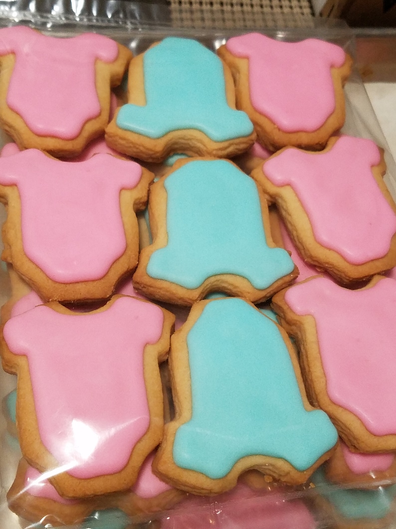 May include: A close-up of a variety of sugar cookies decorated with blue and pink icing in the shape of onesies. The cookies are arranged in a plastic bag.