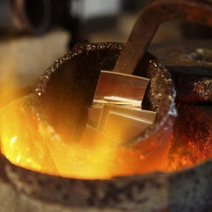 K&ouml;nnte beinhalten: Nahaufnahme eines Metalltiegels mit geschmolzenem Metall darin. Das Metall gl&uuml;ht orange und wird mit einem Metallwerkzeug ger&uuml;hrt. Der Tiegel steht auf einem Bett aus gl&uuml;henden Kohlen.