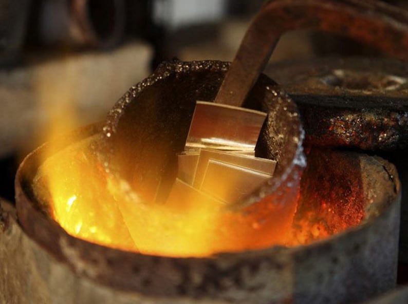 K&ouml;nnte beinhalten: Nahaufnahme eines Metalltiegels mit geschmolzenem Metall darin. Das Metall gl&uuml;ht orange und wird mit einem Metallwerkzeug ger&uuml;hrt. Der Tiegel steht auf einem Bett aus gl&uuml;henden Kohlen.