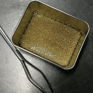 K&ouml;nnte beinhalten: Ein kleiner, rechteckiger Metallbeh&auml;lter gef&uuml;llt mit winzigen, gl&auml;nzenden goldenen Perlen. Der Beh&auml;lter steht auf einer grauen Oberfl&auml;che neben einer Metallpinzette.