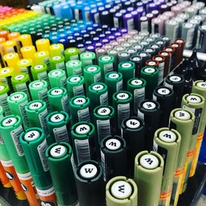 May include: A close-up view of a variety of green, blue, yellow, and black markers. The markers are arranged in rows and columns, and the caps are all facing the camera. The markers are labeled with the letter "W" and a number.
