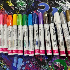 May include: A collection of Sharpie oil-based paint markers in various colors, including purple, pink, orange, yellow, green, blue, and brown. The markers have white barrels with the Sharpie logo and color names. The caps match the ink color.