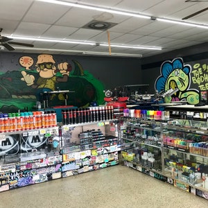May include: A shop interior with a variety of spray paint cans and markers on shelves. The walls are decorated with graffiti art, including a cartoon character and the words "The Bodhi Tree".