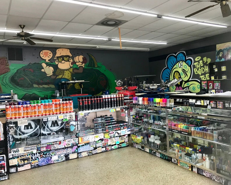 May include: A shop interior with a variety of spray paint cans and markers on shelves. The walls are decorated with graffiti art, including a cartoon character and the words "The Bodhi Tree".