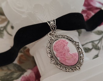 May include: A black velvet choker necklace with a silver cameo pendant featuring a pink profile of a woman. The cameo is surrounded by an ornate silver frame.
