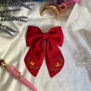 Puede incluir: Una pajarita roja con medias lunas y flores bordadas. La pajarita está sobre un fondo de tela blanca.