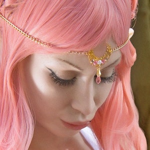 Könnte beinhalten: Eine Frau mit langen, pinkfarbenen Haaren, die zu Zöpfen geflochten sind und einem goldenen Stirnband mit rosafarbenen Edelsteinen. Sie trägt ein weißes Tanktop und hat ein Halbmond-Tattoo auf ihrer Schulter.