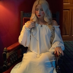 May include: A woman with long blonde hair sits in a dark wood chair with red velvet upholstery. She is wearing a white off-the-shoulder dress with long sleeves and a silver necklace. The background is a dark red wall.