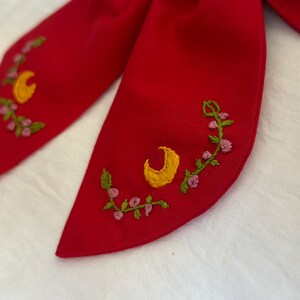 K&ouml;nnte beinhalten: Roter Stoffkragen mit gestickten Halbmonden und Blumendesigns in Gelb, Gr&uuml;n und Rosa.