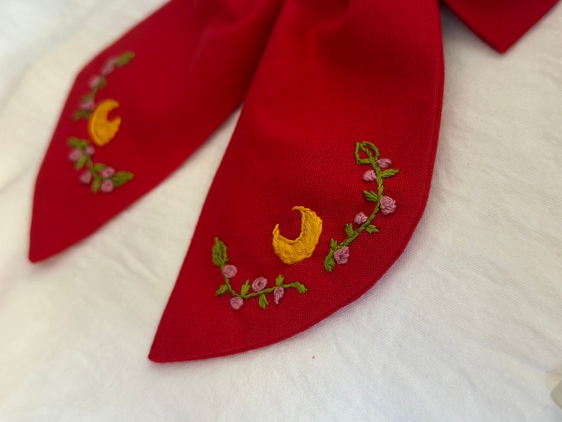 K&ouml;nnte beinhalten: Roter Stoffkragen mit gestickten Halbmonden und Blumendesigns in Gelb, Gr&uuml;n und Rosa.